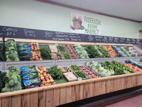 A vibrant display of fresh vegetables and fruits at Rosebush Farm Market, with prices listed on chalkboards above.