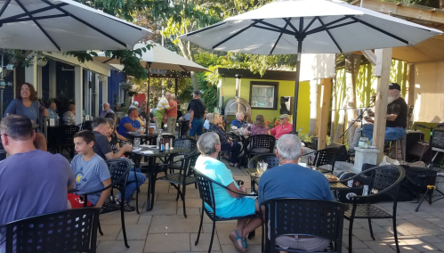 A lively outdoor café scene with people seated at tables, enjoying drinks and live music under large umbrellas.