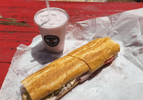 A toasted sandwich with various fillings next to a cup of pink milkshake on a red wooden surface.