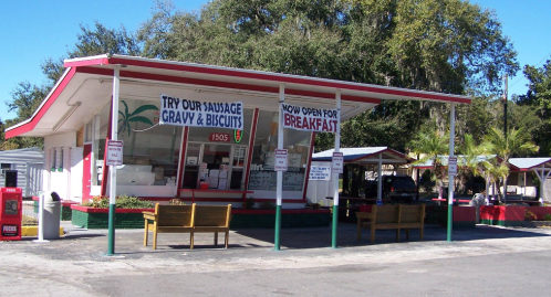 A small diner with a red and white exterior, featuring signs for breakfast and sausage gravy, surrounded by trees.