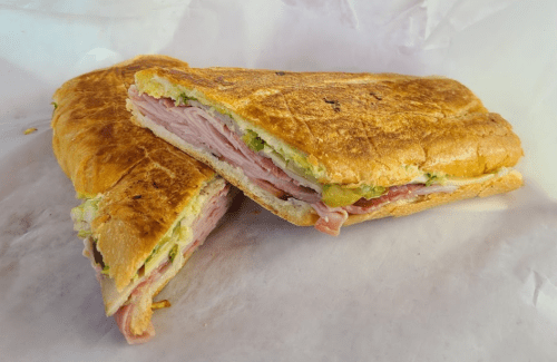 A toasted sandwich filled with ham, lettuce, and pickles, cut in half and resting on white paper.