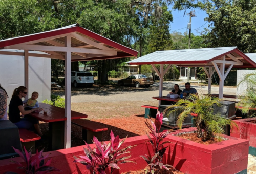 Outdoor seating area with picnic tables, surrounded by plants, where families are enjoying a meal on a sunny day.