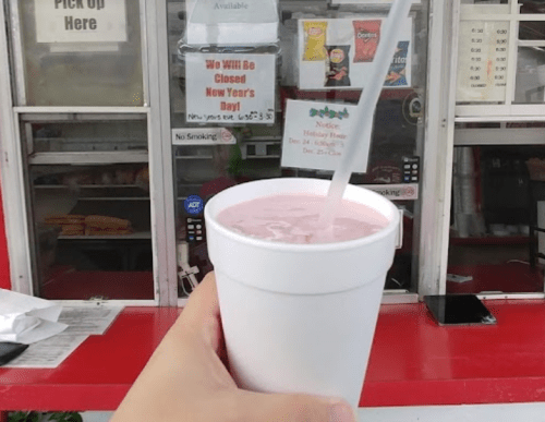 A hand holds a white cup with a pink drink in front of a food stand with a closed sign and menu.
