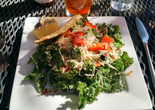 A fresh kale salad topped with diced tomatoes, cheese, and a lemon wedge, served on a white plate.