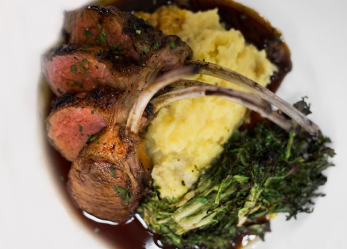 A plate featuring grilled lamb chops, creamy mashed potatoes, and sautéed greens, drizzled with sauce.