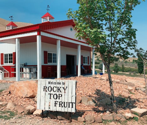 A white building with red accents and a sign reading "Welcome to Rocky Top Fruit" surrounded by trees and rocks.