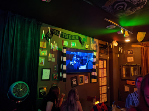 A cozy bar with green lighting, featuring a TV showing a scene from a movie, surrounded by eclectic wall decor.