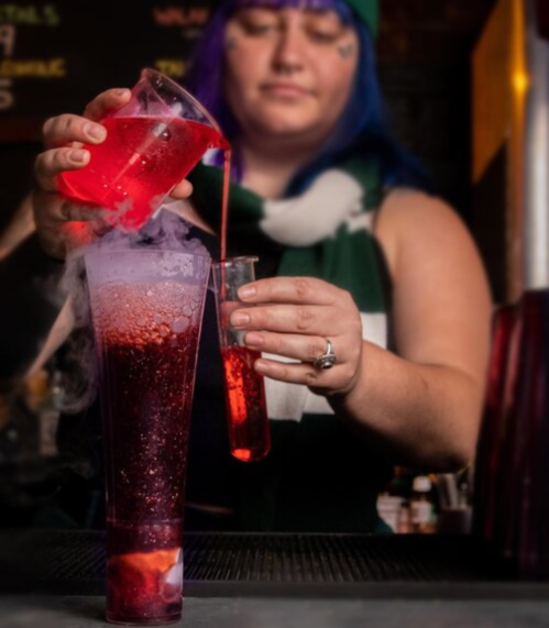 A bartender pours a colorful drink into a tall glass, creating a smoky effect with vibrant red liquid.