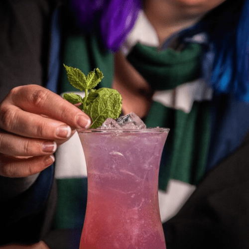 A hand garnishes a pink drink with mint leaves, ice visible in the glass, against a blurred background.