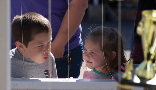 Two children, a boy and a girl, are playfully interacting through a window, with a trophy visible in the foreground.