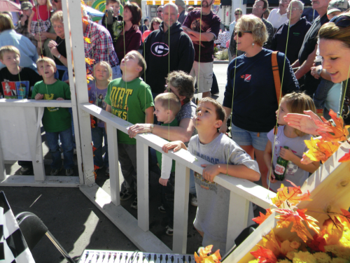 A group of children and adults watch an event at a fair, with colorful decorations and a festive atmosphere.
