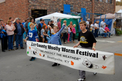 Children hold a banner for the Lee County Woolly Worm Festival, with a crowd and tents in the background.
