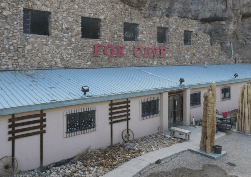Exterior view of Fox Cave, a stone and stucco building with a metal roof, surrounded by rocky terrain and cacti.