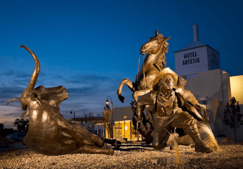 Bronze sculpture of a cowboy on horseback, with a steer lying down, set against a twilight sky and a hotel backdrop.