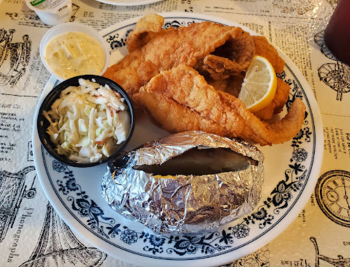 A plate with two pieces of fried fish, a lemon wedge, coleslaw, and a baked potato wrapped in foil.