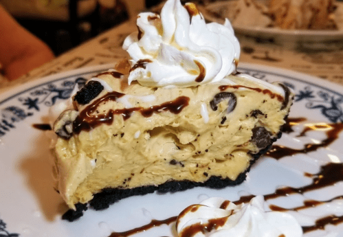 A slice of creamy peanut butter pie topped with whipped cream and chocolate drizzle on a blue-patterned plate.