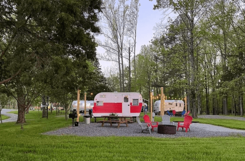 A scenic campsite with vintage trailers, surrounded by trees and a fire pit with colorful chairs.