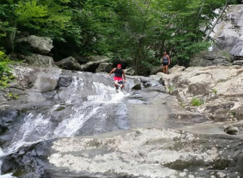 A person in red shorts walks down a rocky stream, while another person stands nearby among lush greenery.