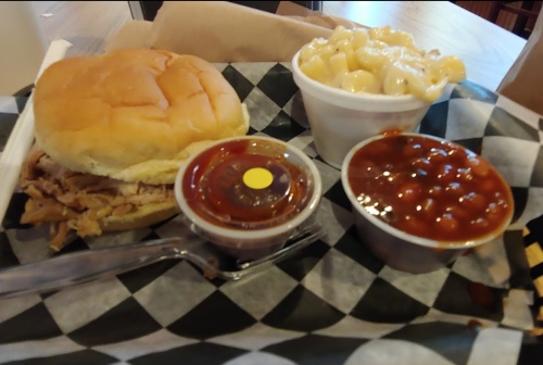 A tray with a pulled pork sandwich, macaroni and cheese, baked beans, and a small cup of barbecue sauce.