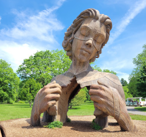 A large wooden sculpture of a woman with closed eyes, holding her chest, surrounded by greenery and blue skies.