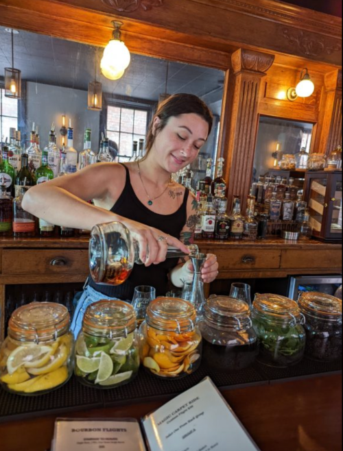 A bartender pours a drink while surrounded by jars of fresh fruit at a wooden bar.