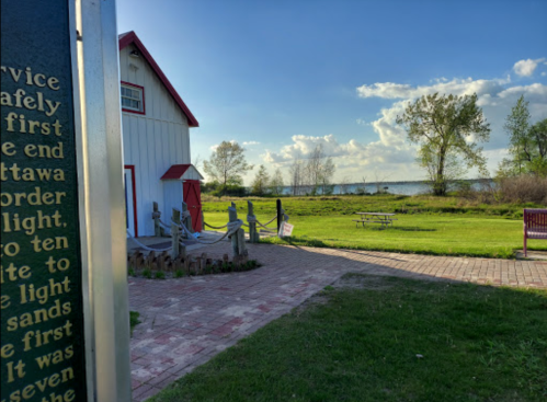 A scenic view of a grassy area with a path, a red-roofed building, and a lake under a blue sky with clouds.