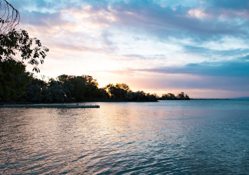 A serene lake at sunset, with colorful clouds reflecting on the water and trees lining the shore.