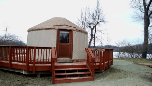 A tan yurt on a wooden deck by a lake, surrounded by bare trees and a cloudy sky.