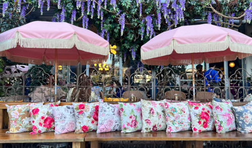 A cozy outdoor seating area with pink umbrellas and floral cushions, surrounded by hanging purple flowers.