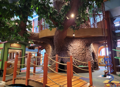 A large, textured tree structure inside a play area, with a wooden bridge and colorful play equipment nearby.