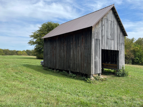 A weathered wooden barn stands in a grassy field under a blue sky with scattered clouds.