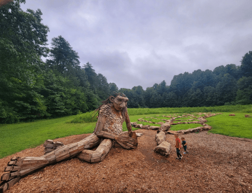 A large wooden sculpture of a seated creature in a forested area, with two children exploring nearby.