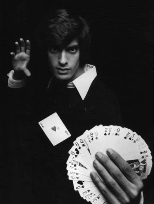 A magician holds a fan of playing cards, showcasing an ace of hearts, with a mysterious expression.