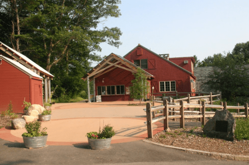 A red building with a sloped roof surrounded by greenery and a gravel path, featuring planters and a stone marker.