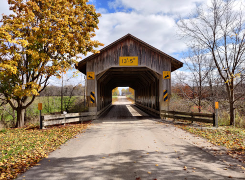 A wooden covered bridge with a low clearance sign, surrounded by autumn trees and a clear blue sky.