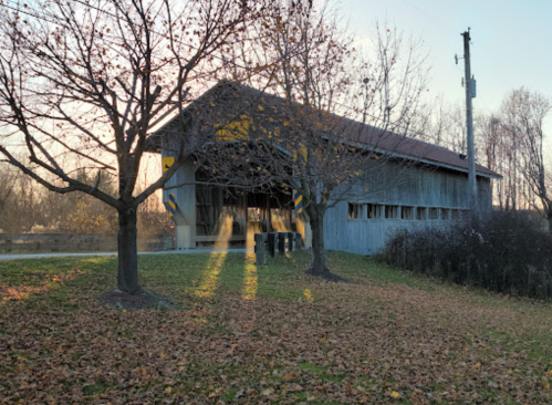A covered bridge surrounded by trees and fallen leaves, with sunlight streaming through its openings.