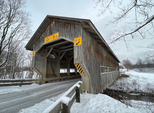 A snow-covered wooden covered bridge with a low clearance sign, surrounded by bare trees and a winter landscape.