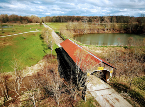 Aerial view of a covered bridge over a road, surrounded by trees and a pond under a partly cloudy sky.