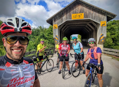 A group of six cyclists poses in front of a covered bridge, smiling and wearing colorful cycling gear.