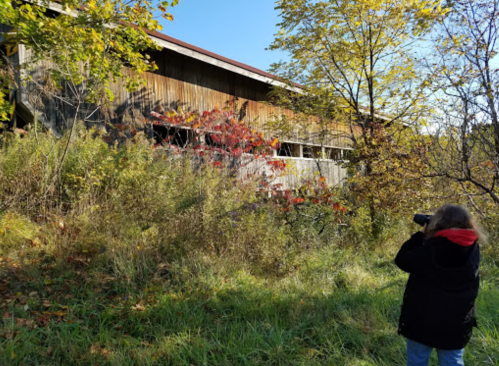 A person in a black jacket takes a photo of an old wooden building surrounded by greenery and autumn foliage.