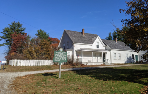 A white historic house with a porch, surrounded by trees and a grassy area, under a clear blue sky.