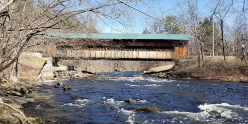 A wooden covered bridge spans a flowing river, surrounded by bare trees and a clear blue sky.