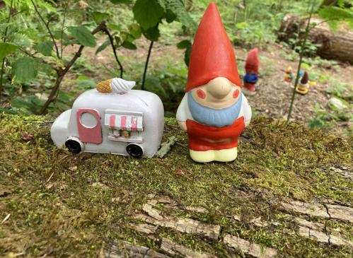 A colorful gnome stands next to a small, whimsical food truck in a forest setting, surrounded by greenery.