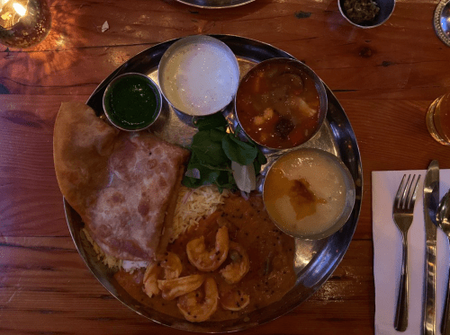 A silver platter with rice, shrimp, a flatbread, yogurt, green sauce, and a bowl of soup, set on a wooden table.