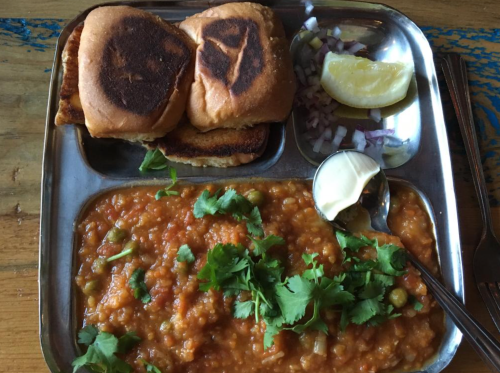 A metal plate with two toasted buns, a bowl of spicy lentil dish topped with cilantro, onions, and a lemon wedge.
