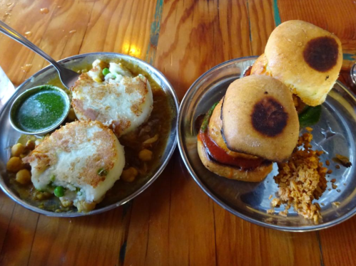 Two metal plates with Indian street food: one with chickpea curry and fried dumplings, the other with sliders and crispy rice.