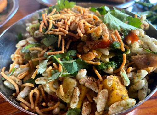 A close-up of a colorful snack mix with crispy noodles, fresh herbs, and diced fruits in a metal bowl.