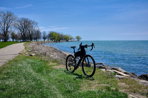 A black bicycle parked on a grassy path by a lake, with trees and a clear blue sky in the background.