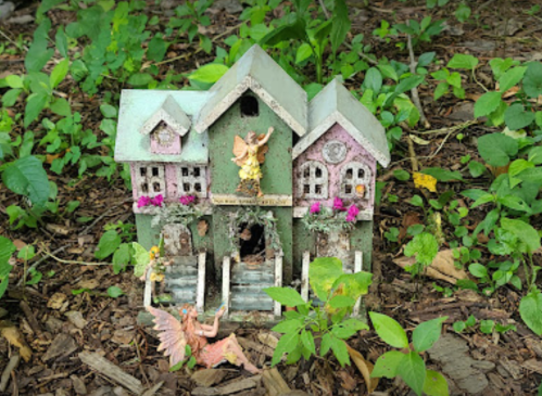 A small, whimsical fairy house surrounded by greenery, with a fairy figure resting nearby.