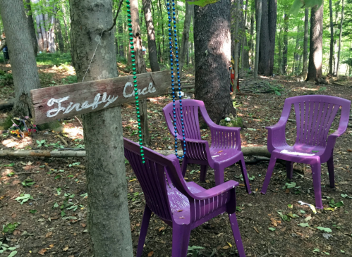 A wooden sign reading "Firefly Circle" hangs on a tree near four purple chairs in a wooded area.